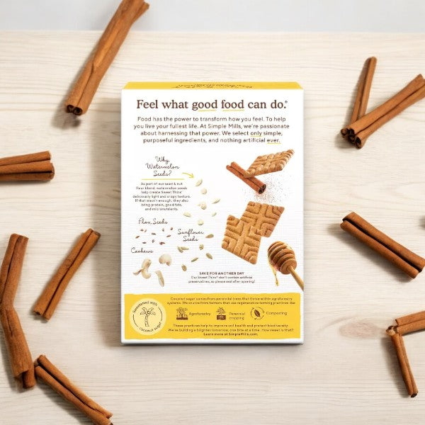 A box of Simple Mills Honey Cinnamon Sweet Thins is surrounded by cinnamon sticks, emphasizing the delectable Honey Cinnamon flavor. The back of the box displays product information alongside a photo showcasing these delicious snacks made with Seed & Nut Flour.