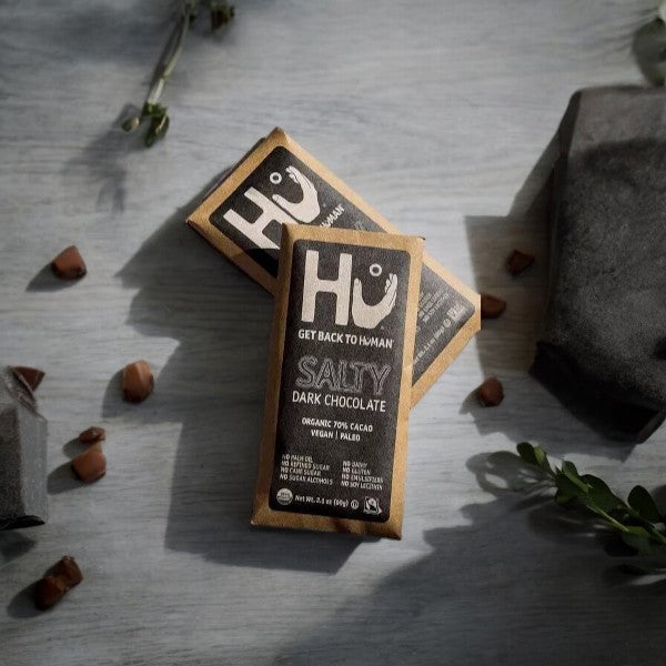 Two Hu Salty Dark Chocolate 70% Cacao bars rest on a surface with scattered hazelnuts and greenery. The packaging emphasizes its organic ingredients, 70% cacao content, and vegan and paleo certifications.