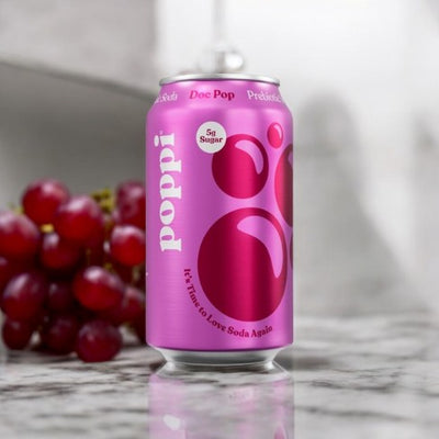 A can of Poppi: Doc Pop Prebiotic Soda in a pink design is placed on a marble surface next to a small bunch of red grapes, offering a healthy alternative as a low-calorie beverage.