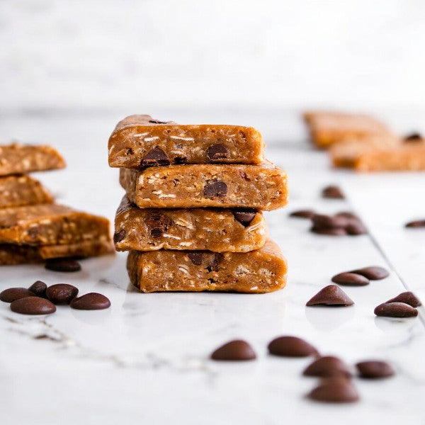 A stack of four GoMacro Oatmeal Chocolate Chip MacroBars from GoMacro, each 2.3 oz and gluten-free, sits on a white marble surface, surrounded by scattered chocolate chips.