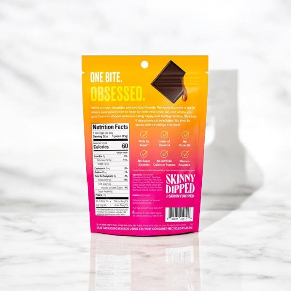 The back view of a SkinnyDipped Dark Chocolate Salted Caramel Bite Candy Bag (3 oz) package showcases the "One Bite. Obsessed." slogan, nutrition facts, ingredients, and brand details against a marble background. This gluten-free and keto-friendly treat from Skinny Dipped is as delightful as it is guilt-free.