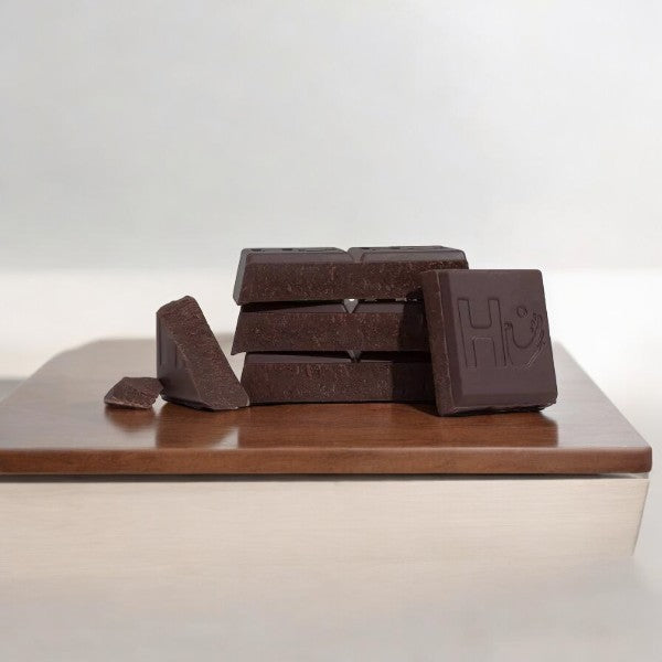 A stack of Hu Simple Dark Chocolate 70% Cacao Candy bars on a wooden surface, with one bar featuring the letters "Hu" engraved on it. Some pieces, sweetened with organic unrefined coconut sugar and boasting 70% cacao content, are broken off and placed nearby.