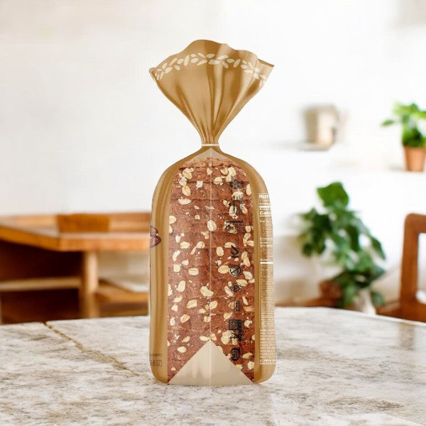 A loaf of Silver Hills Bakery Vegan The Big 16 Sprouted Grain Bread in a clear and brown plastic bag sits on a marble countertop, with a blurred kitchen in the background.