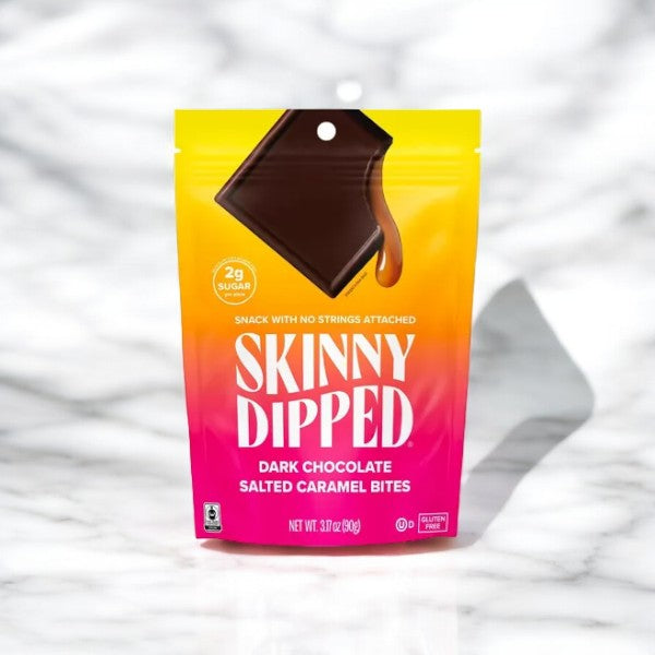 A bag of SkinnyDipped Dark Chocolate Salted Caramel Bite Candy Bag 3 oz standing upright. The packaging is a gradient from yellow to pink, featuring an image of a chocolate square and text detailing product features. These keto-friendly treats from Skinny Dipped are also gluten-free, making them a delicious and mindful choice.