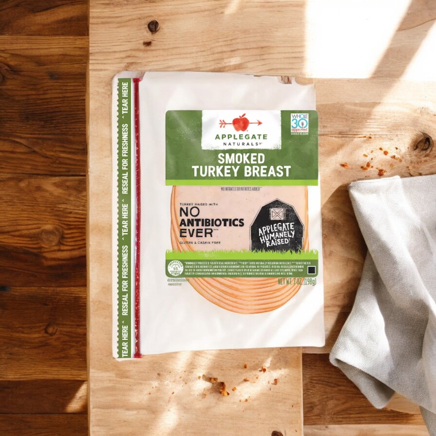 A package of Applegate Natural Smoked Turkey Breast, 7 oz - 1 Count, labeled as Fully Cooked and Antibiotic-Free, sits on a wooden surface with a cloth nearby. The packaging highlights "No Antibiotics Ever" and "Applegate Humanely Raised".