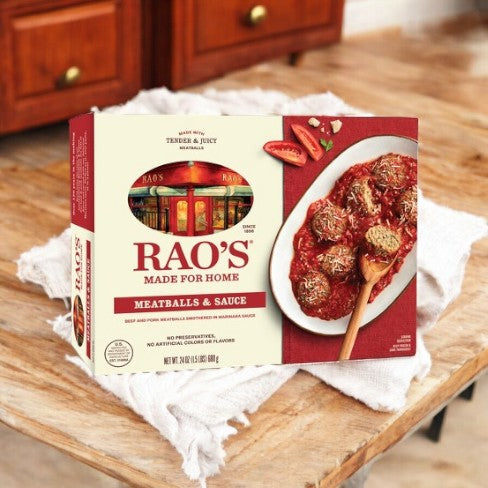 A box of Easy Lunches Rao's Made For Home Family Size Frozen Meatballs and Sauce - 24 oz is placed on a white cloth on a wooden surface, featuring a picture of meatballs in marinara sauce on the packaging.