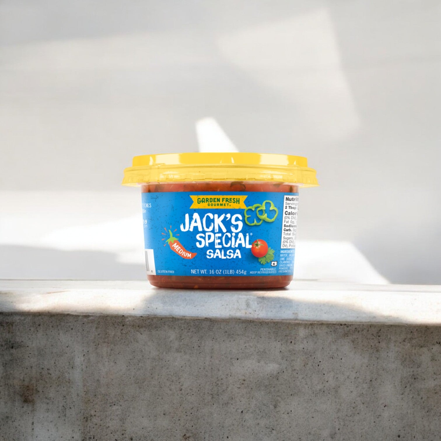 A 16 oz. container of Garden Fresh Gourmet Jack's Special Medium Salsa from the Easy Lunches brand, featuring a yellow lid, is placed on a concrete surface.