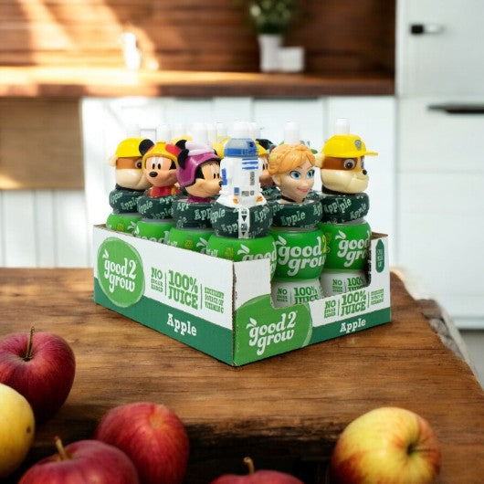 A display of Easy Lunches' Good2Grow Apple Juice bottles, featuring collectible character tops such as Mickey Mouse and various other cartoon figures, is placed on a wooden table with fresh apples. Each 6-fluid ounce bottle contains 100% pure apple juice, making it both a fun and healthy choice for kids. Available in cases of 12.