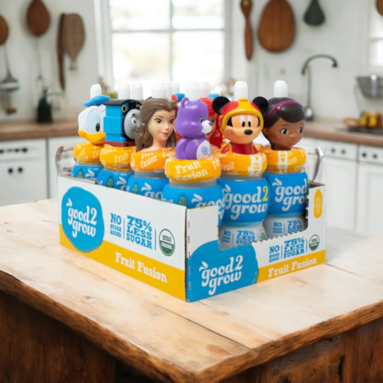 A box of Easy Lunches' Good2grow Single Serve Fruit Fusion healthy kids drinks, each featuring varying character-themed tops, is displayed on a kitchen island with white cabinets and wooden accents in the background.