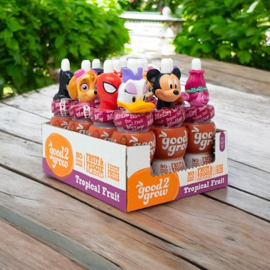 A display box of Easy Lunches Good2grow Single Serve Tropical Fruit Medley Juice bottles, each 6 fluid ounces and topped with a varying collectible character top, is placed on a wooden table outdoors.