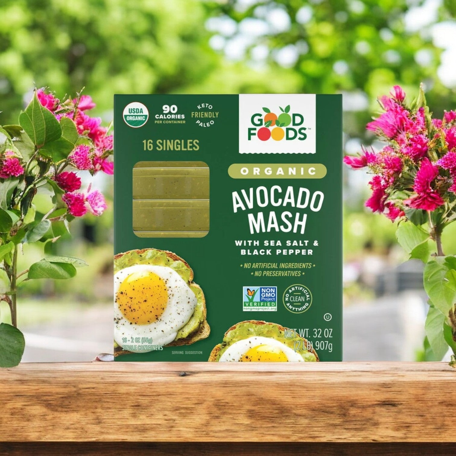 A pack of Good Foods Organic Avocado Mash with Sea Salt & Black Pepper, featuring 2 oz portions in a 16-count pack, sits on a table outdoors surrounded by pink flowers, offering convenient portions for any occasion.