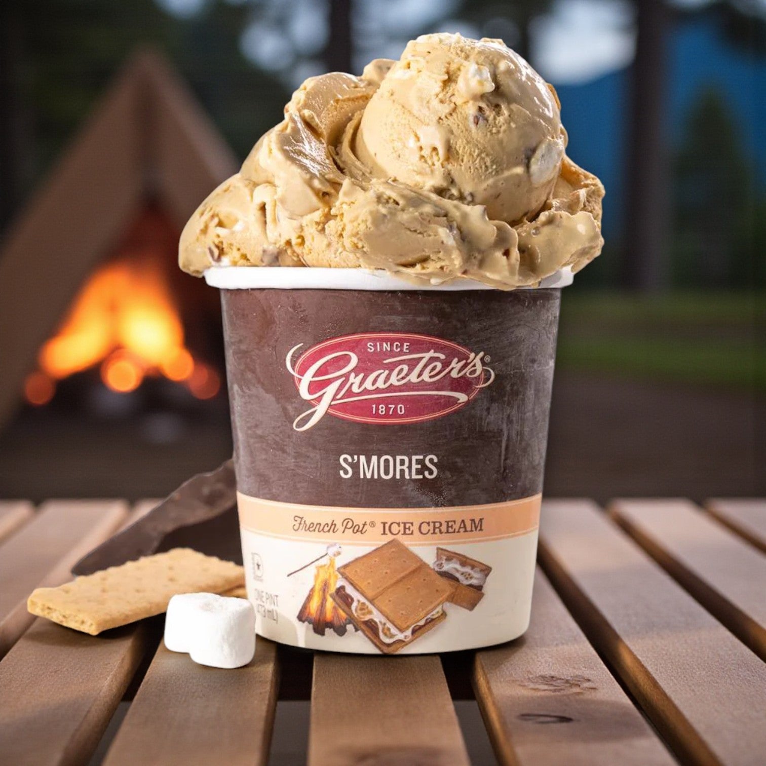 A Graeter’s Craft Ice Cream Combo Pack – 6 Pints sits on a wooden table with marshmallow, graham cracker, and chocolate beside it; a campfire and tent are blurred in the background.