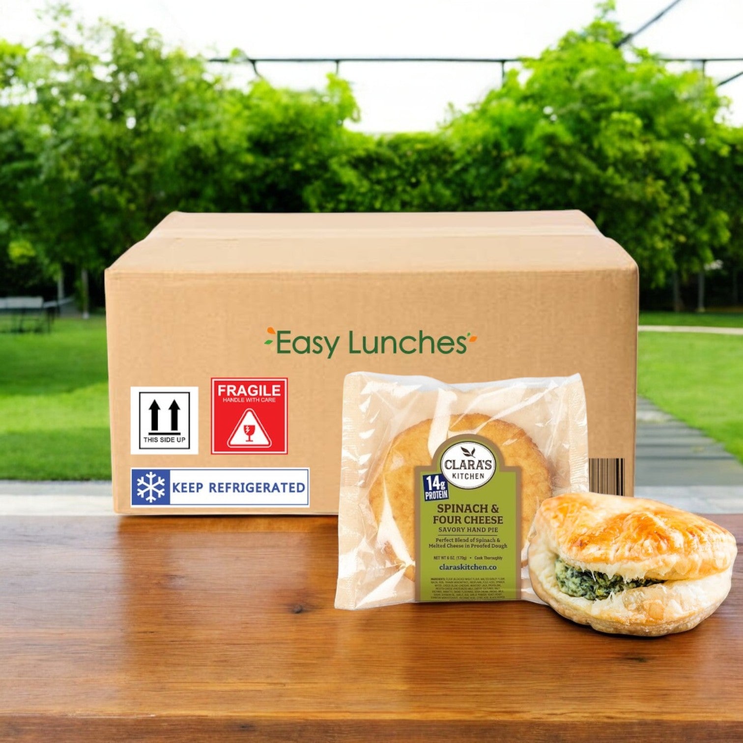 A cardboard box labeled "Easy Lunches" sits on a wooden table outdoors, showcasing Clara's Kitchen Four Cheese Spinach Handpie, 6 oz.—vegetarian-friendly and gourmet comfort food—with another croissant to the side.