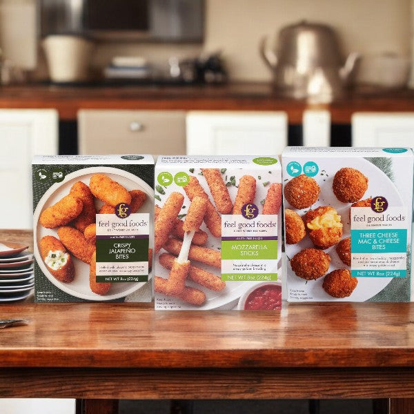 Three boxes of Easy Lunches' Feel Good Variety Pack! frozen snacks are displayed on a wooden kitchen counter, featuring Crispy Jalapeño Bites, Mozzarella Sticks, and Three Cheese Mac & Cheese Bites.