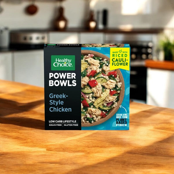 A box of Healthy Choice Power Bowls labeled "Greek-Style Chicken" sits on a wooden kitchen counter. The packaging highlights that it is made with riced cauliflower, low carb, grain-free, and gluten-free—perfect for those seeking nutritious microwaveable meals within the Healthy Choice Power Bowl Variety Pack, which includes 2 Korean Style Chicken, 2 Adobo Chicken, 2 Chicken Marinara, and 2 Greek Style Chicken for a total of 8 delicious Power Bowls.