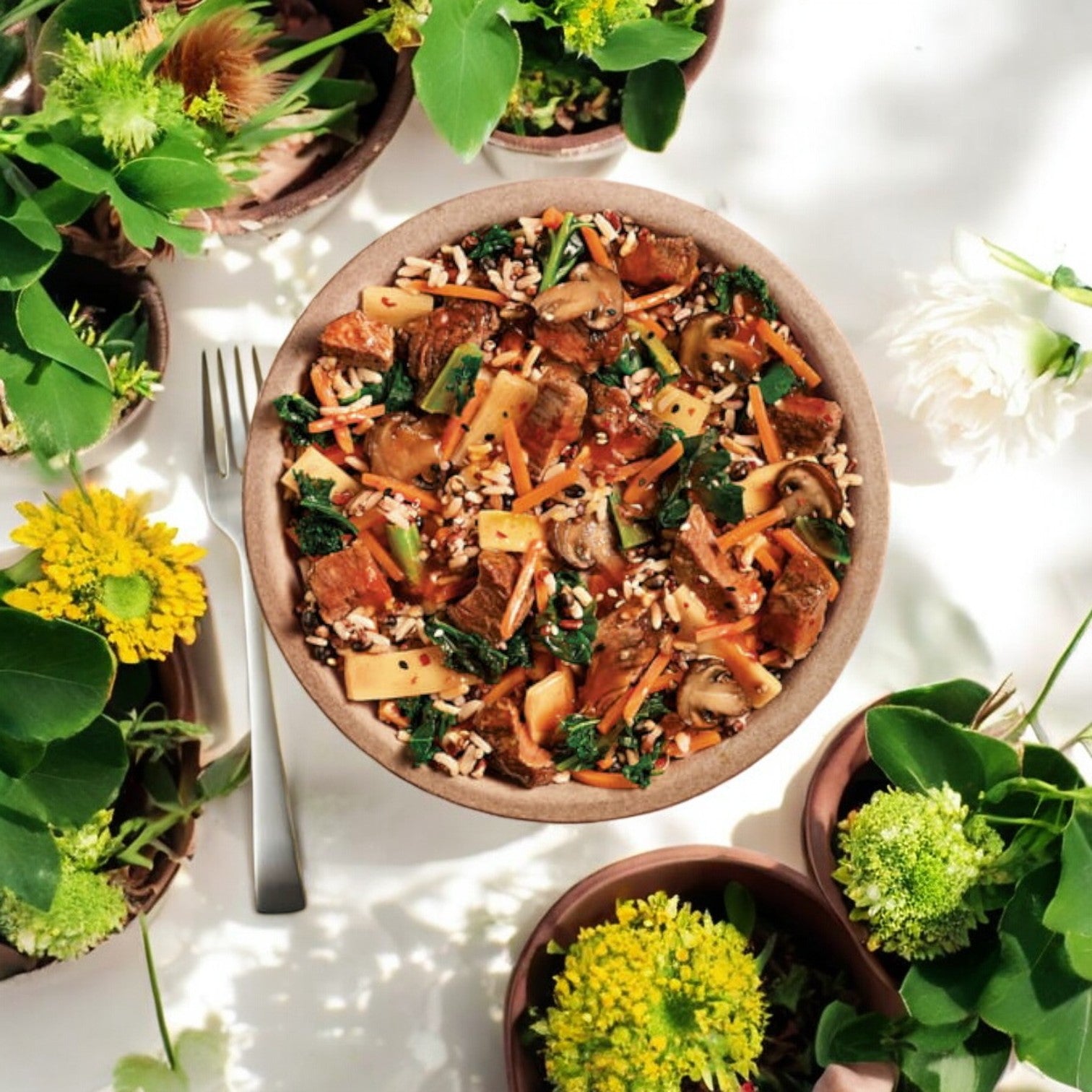 A bowl of stir-fried vegetables and tofu on a white table, surrounded by various green and yellow potted plants, resembles a Healthy Choice Power Bowls Korean Beef. A fork lies next to the bowl.