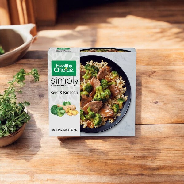 A Healthy Choice Simply Steamers Beef & Broccoli, 10 oz. - 1 Count package is placed on a wooden surface next to a plant pot. The image showcases the box and the meal inside, highlighting its convenient steam-in-bag technology that ensures freshness.