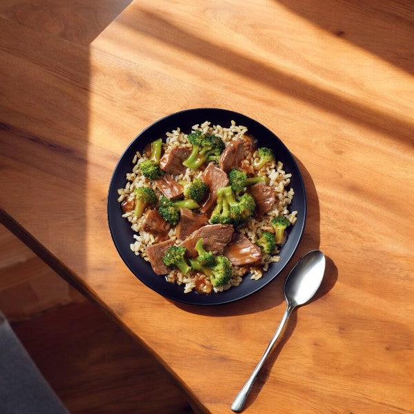 A plate of Healthy Choice Simply Steamers Beef & Broccoli, 10 oz. - 1 Count, is placed on a wooden table with a spoon beside it. Sunlight casts shadows across the surface, highlighting the freshness preserved by steam-in-bag technology.