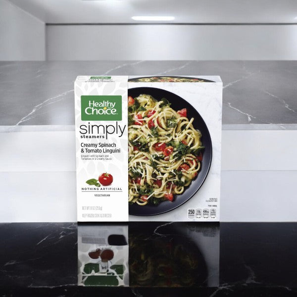 In a modern kitchen setting, a box of Healthy Choice Simply Steamers Creamy Spinach & Tomato Linguini, 9 oz. - 1 Count, is displayed on a reflective black surface alongside an appetizing Healthy Choice Power Bowls featuring riced cauliflower.