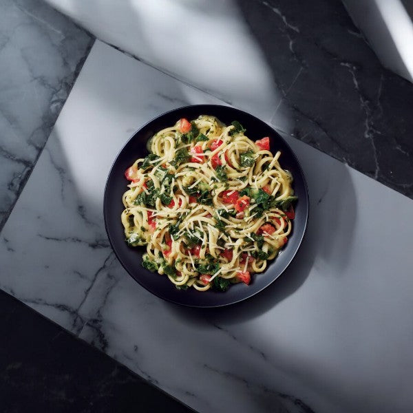 A black bowl filled with Healthy Choice Simply Steamers Creamy Spinach & Tomato Linguini, topped with chopped vegetables, grated cheese, and Greek-style chicken, placed on a black and white marble surface.