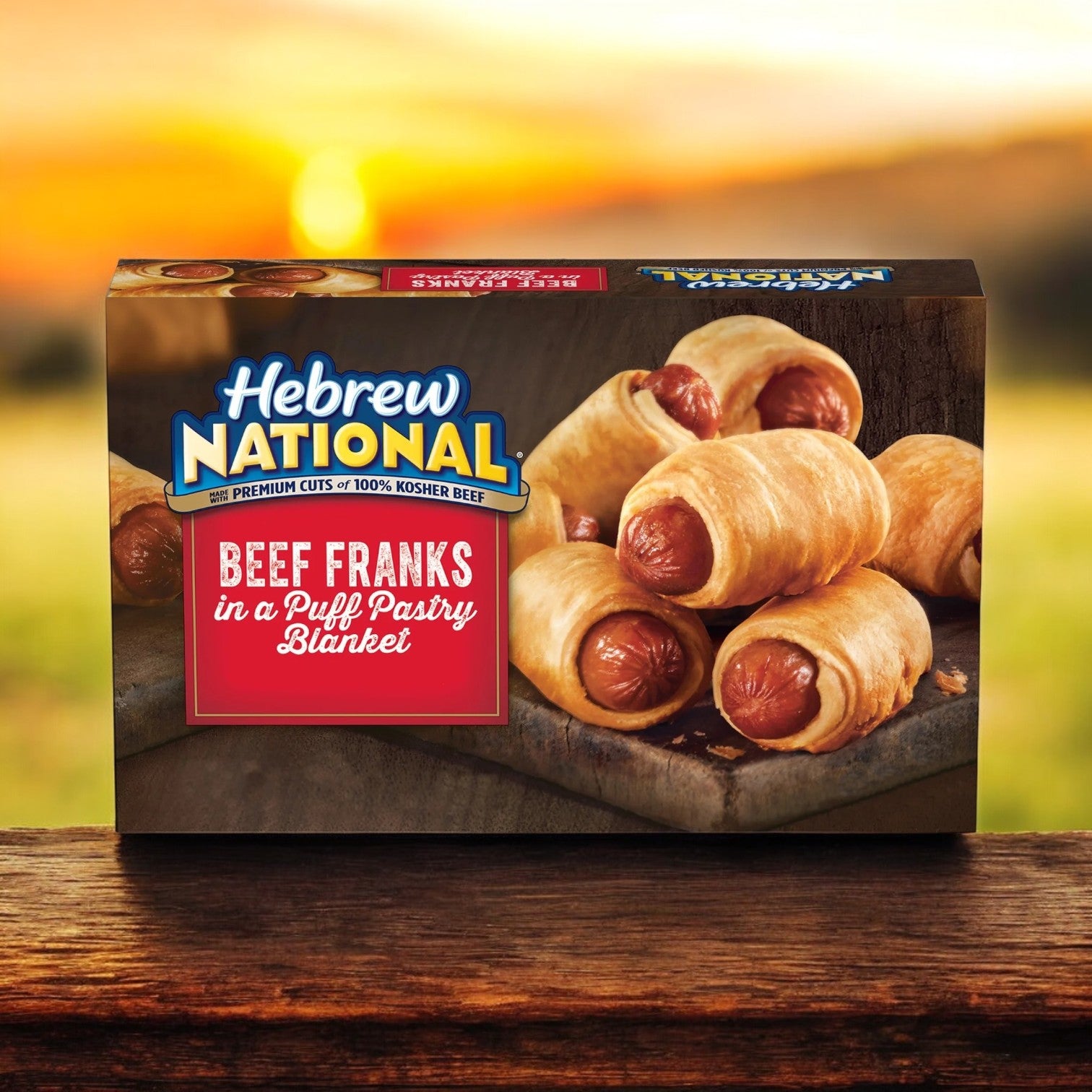 A box of Hebrew National Beef Franks in a Puff Pastry Blanket (18.4 oz, 32 Count Pack) is elegantly displayed on a wooden surface, with a softly blurred nature background enhancing its allure.