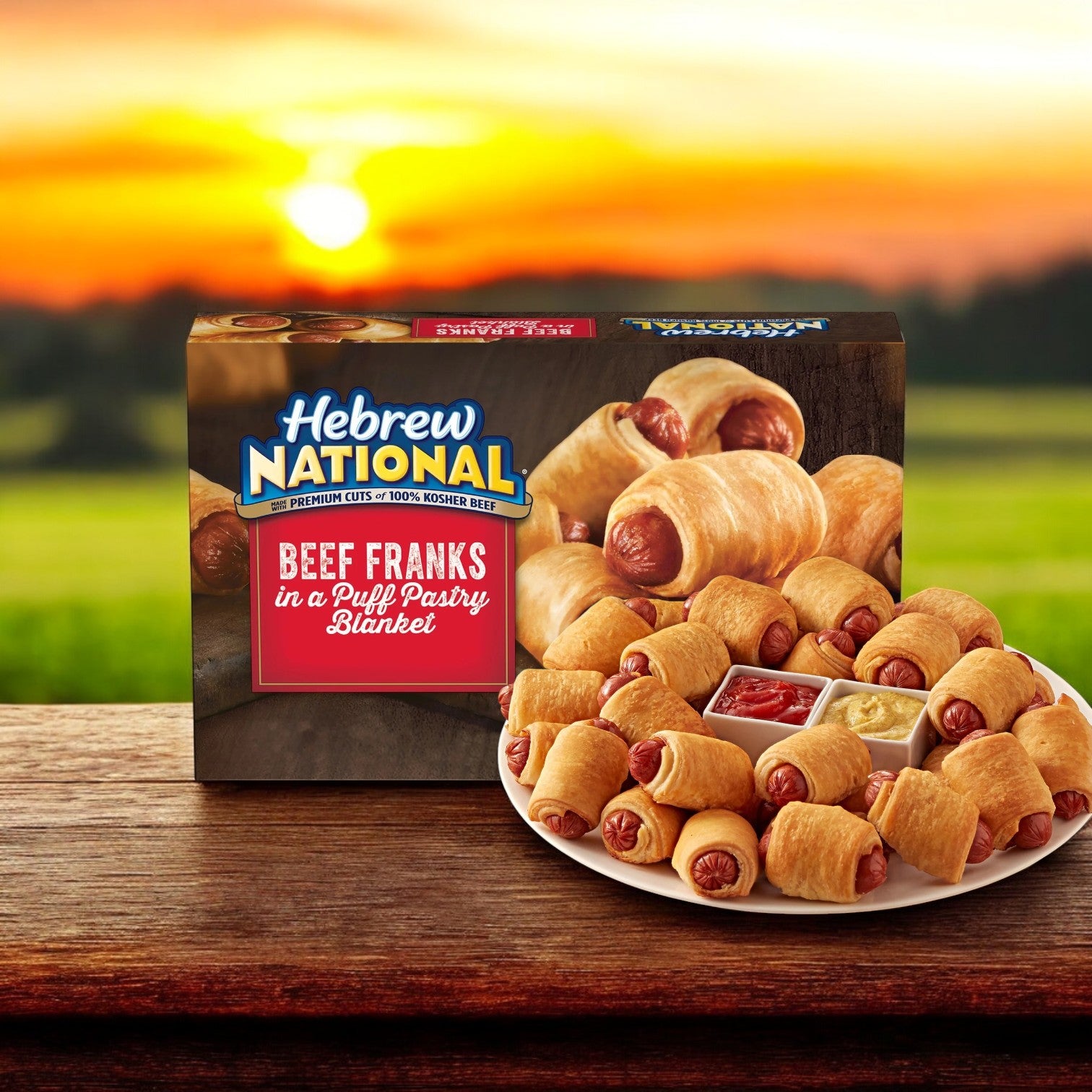 A plate of Hebrew National Beef Franks in a Puff Pastry Blanket (32 count pack, 18.4 oz) sits before its box, set against a backdrop of sunset over a green field.