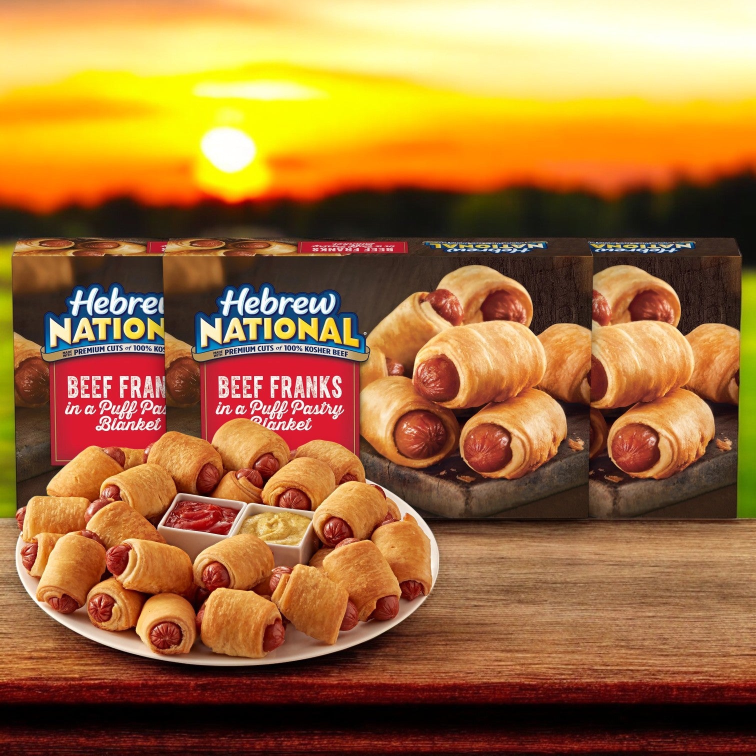 Displayed against a sunset backdrop, Hebrew National Beef Franks in a Puff Pastry Blanket, featuring three 18.4 oz boxes totaling 32 counts, are arranged with a plate of the franks in front.