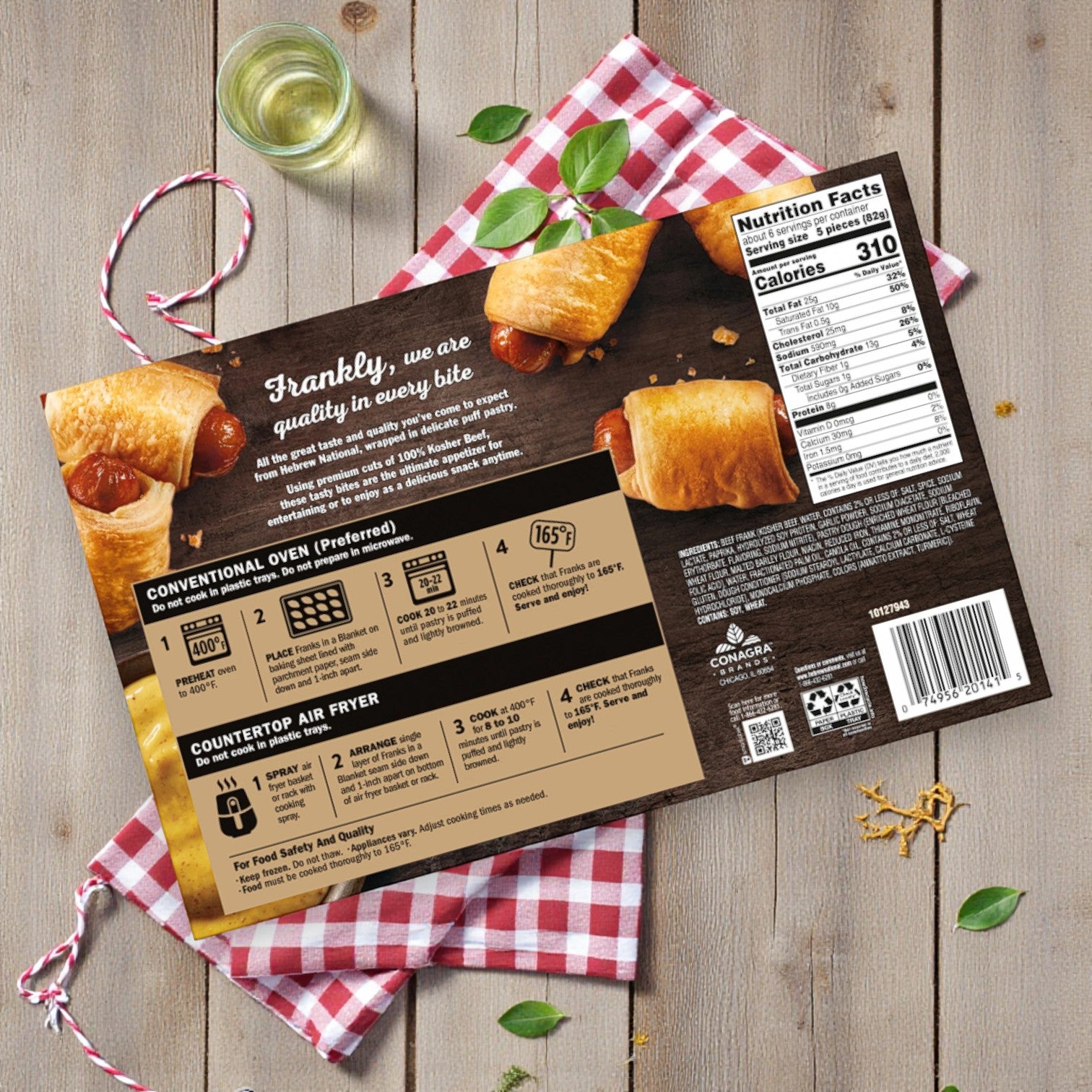 A box of Hebrew National Beef Franks in a Puff Pastry Blanket (3 count, 18.4oz), shown frozen with heating instructions and nutrition facts on a wooden surface with a red and white checkered cloth and basil leaves.