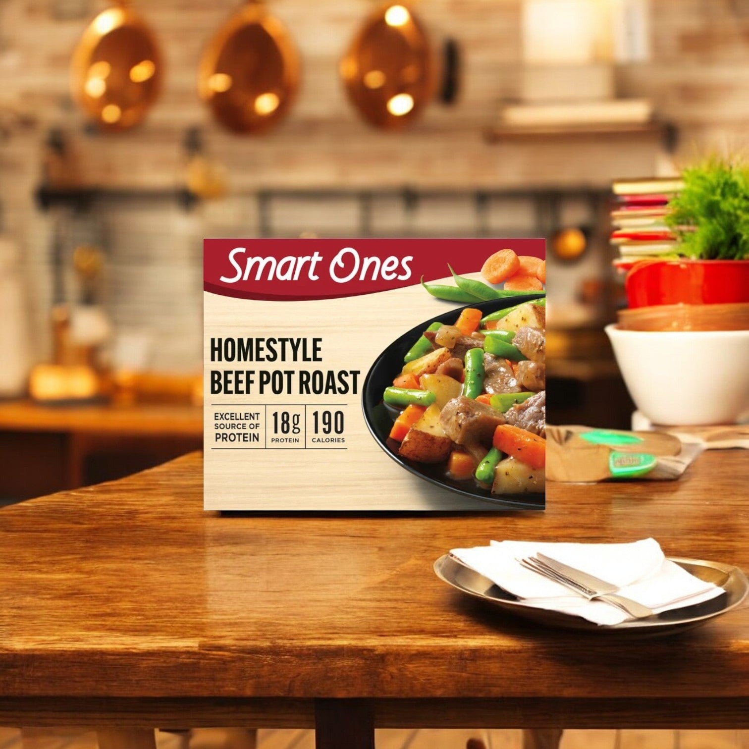 A box of Smart Ones Beef Pot Roast with Carrots, Onions, Potatoes, Green Beans & Gravy is placed on a wooden kitchen table, with a blurred background of kitchen utensils and decor, hinting at a diverse range of flavors from southwestern cuisine to hearty homestyle favorites.