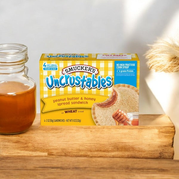 A box of Uncrustable Smuckers Uncrustables Peanut Butter & Honey Spread Sandwiches – 4 Count, 4 Pack- 16 Total Sandwiches sits on a wooden surface next to a small jar of honey. These ready-to-eat sandwiches are perfect for a quick and delicious snack.