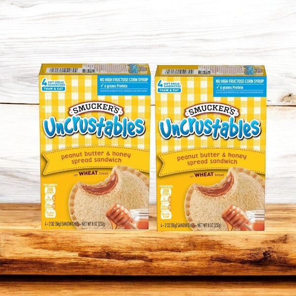 Two packs of Uncrustables Peanut Butter & Honey Spread Sandwiches by Smucker's, each containing four ready-to-eat snacks, sit on a wooden surface. With a total of eight delicious sandwiches, they're perfect for any time you need a quick bite.