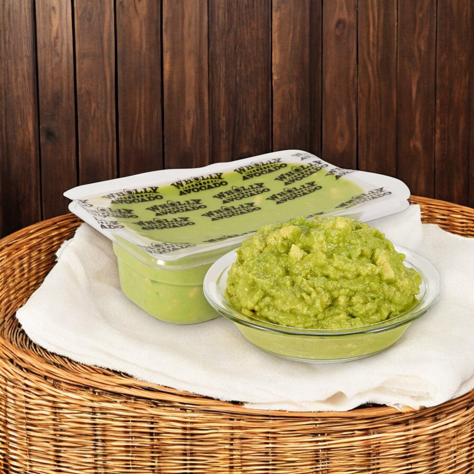 A sealed container of Wholly Guacamole and a bowl of guacamole are placed on a wicker stand against a wooden backdrop.