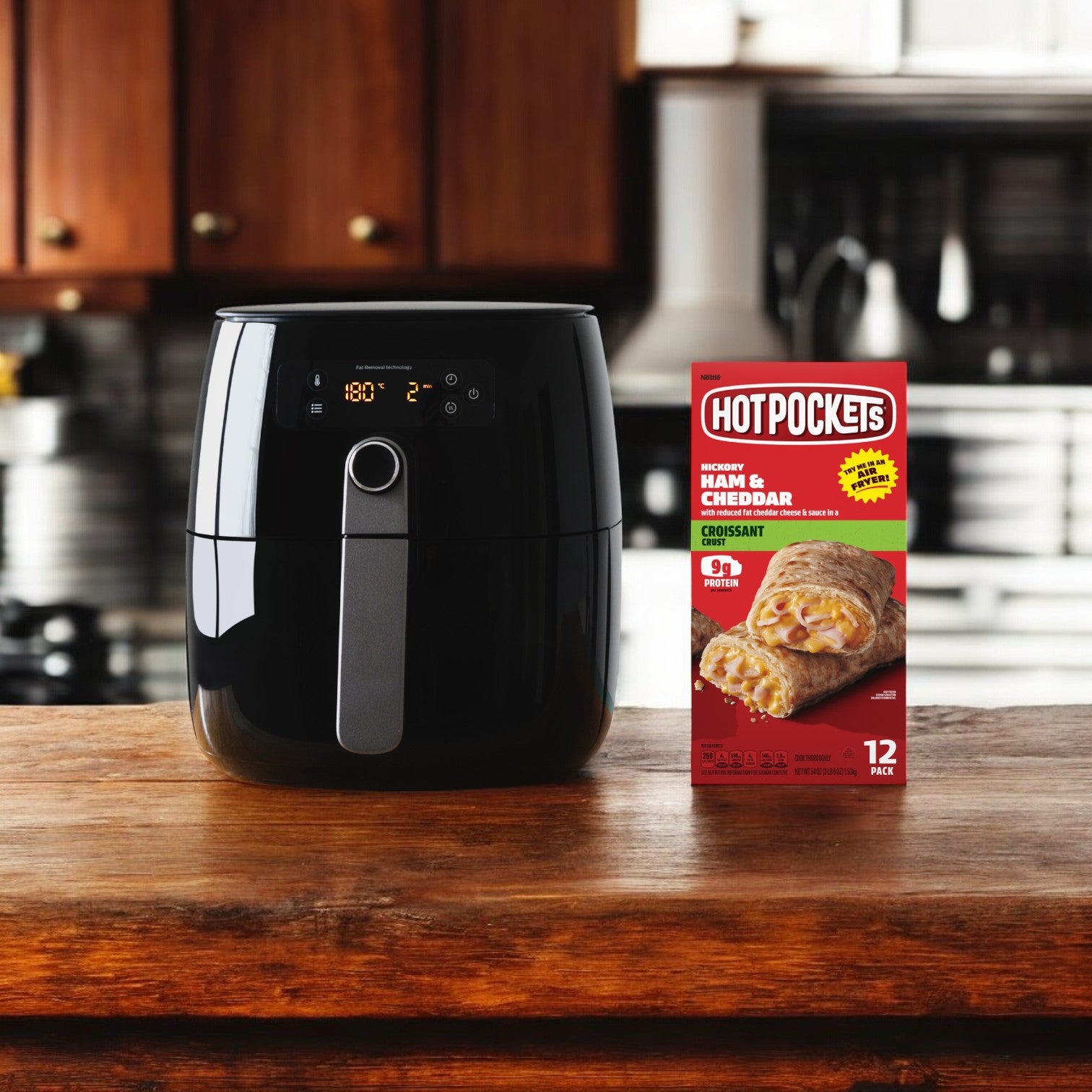 A black air fryer sits on a kitchen counter next to a box of Hot Pockets Frozen Snacks, Ham and Cheddar Croissant Crust, 54 oz. - 1 Pack of 12 Hot Pockets.