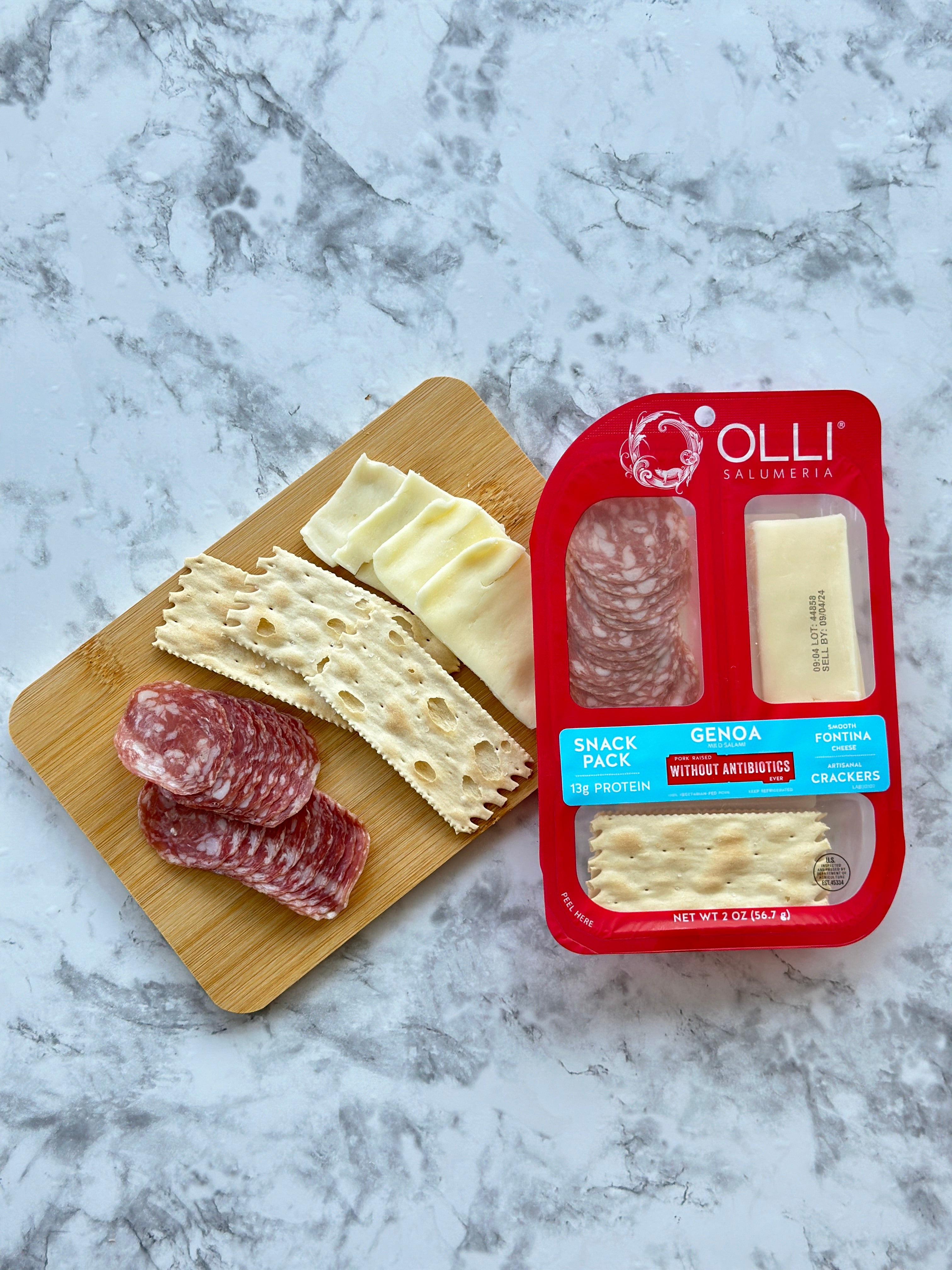 An Olli Salumeria Snack Pack Genoa- 2 oz - 1 Count, showcasing artisanal Italian flavors, is displayed on a marble surface with salami, Fontina cheese, and crackers. Additional Genoa salami, cheese, and crackers from Olli are artfully placed on a small wooden board.