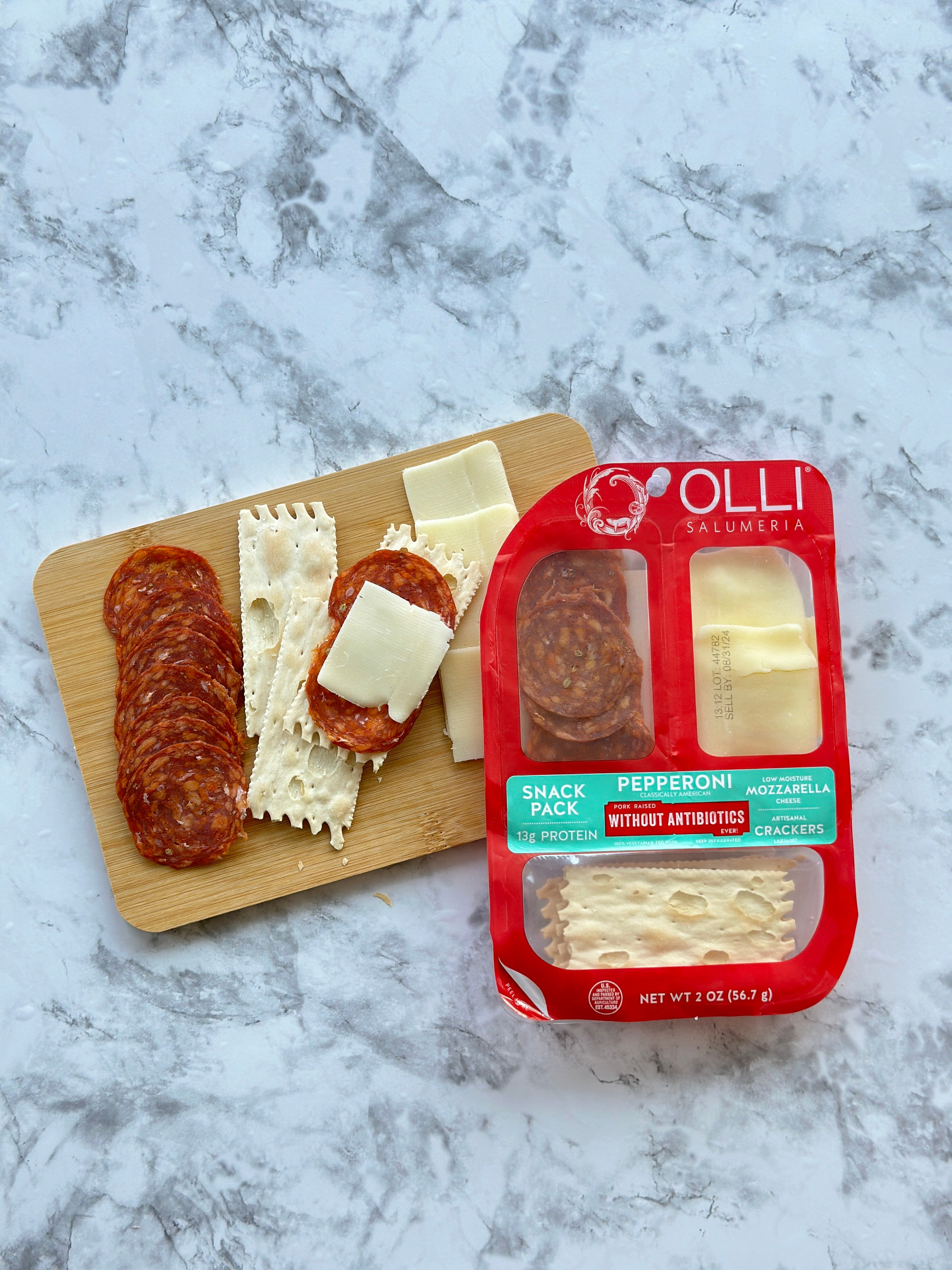 An Olli Salumeria Snack Pack Genoa, including pepperoni, mozzarella, and crackers, is placed next to a wooden board with artisanal Italian flavors of Genoa salami, pepperoni slices, and mozzarella pieces on a marble surface.