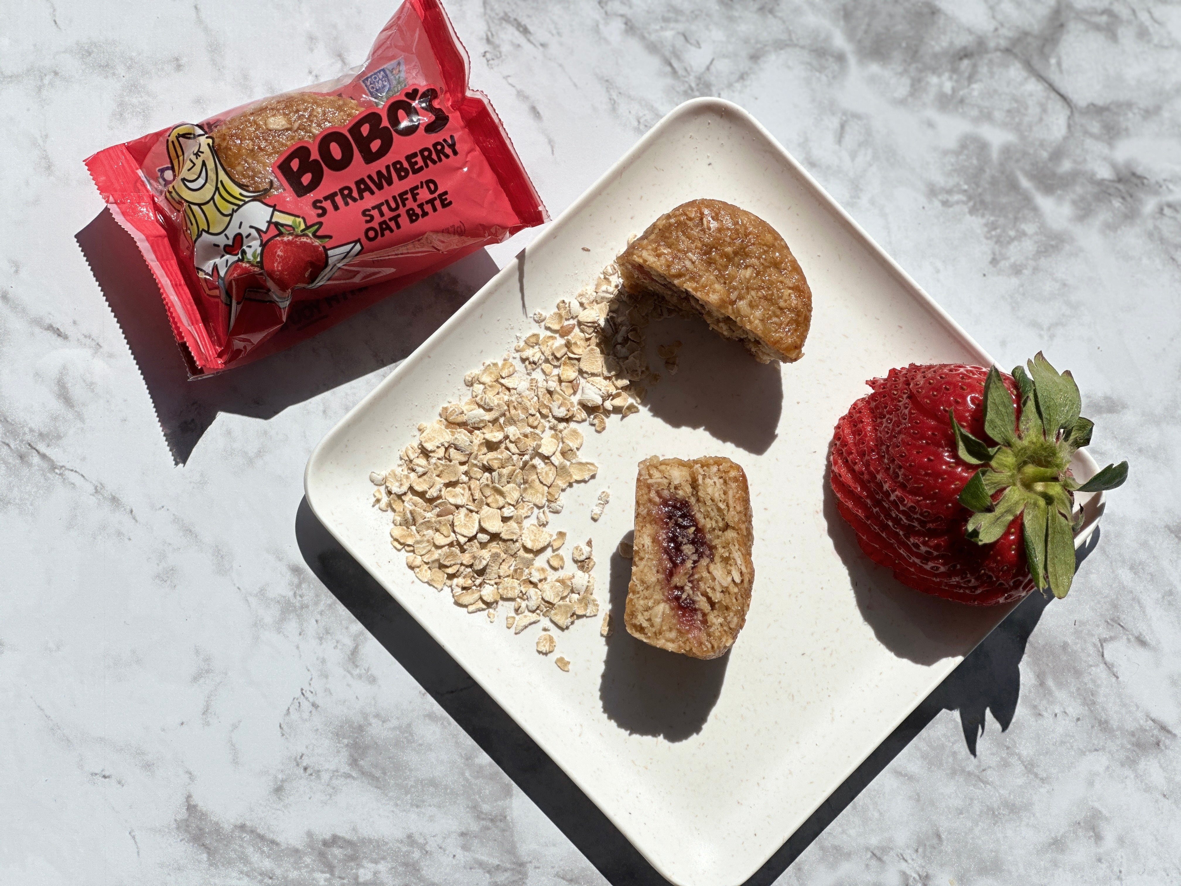 A square white plate featuring a halved strawberry oat bite, scattered oats, and a whole strawberry sits beside an opened package labeled "Bobo's Strawberry Stuff'd Oat Bites." Enjoy these vegan and gluten-free treats from Bobo's any time of day!