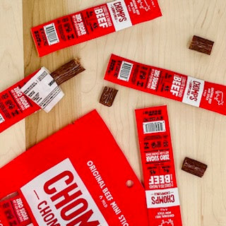 Various opened and unopened Chomp Chomplings Original Beef sticks are scattered on a light wooden surface, showcasing these healthy meat snacks. A red Chomps package is also visible, highlighting their sustainably sourced proteins.