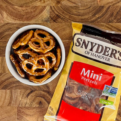 A bowl of bite-sized snacks sits next to an open bag labeled "Snyder's Mini Pretzels" on a wooden surface, showcasing their appeal with natural ingredients.