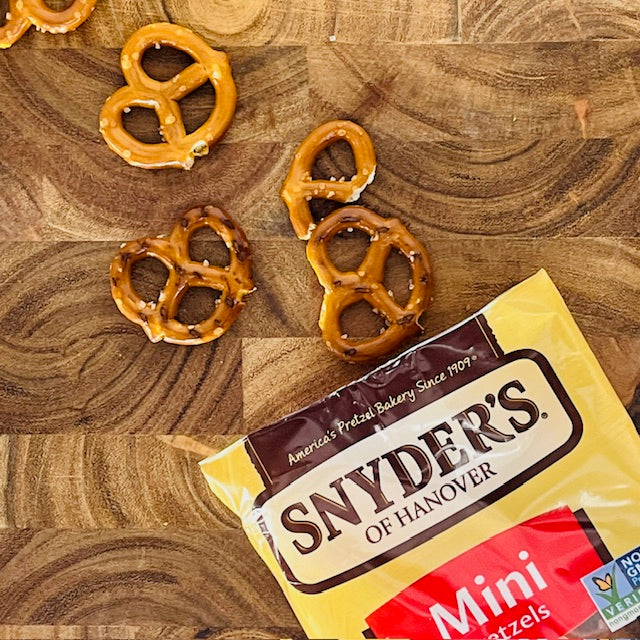 A 1.5 oz. bag of Snyder's of Hanover Mini Pretzels, made with natural ingredients, lies open on a wooden surface with several bite-sized snacks scattered nearby.