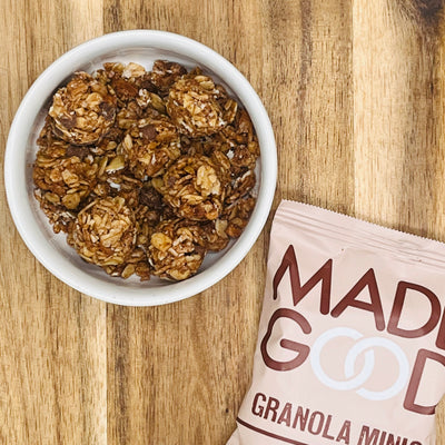 A bowl of granola on a wooden surface. A partially visible bag labeled "MadeGood Chocolate Chip Granola Minis 1-0.85 oz Bag" is placed next to the bowl, highlighting its use of organic ingredients and delightful chocolate chips.