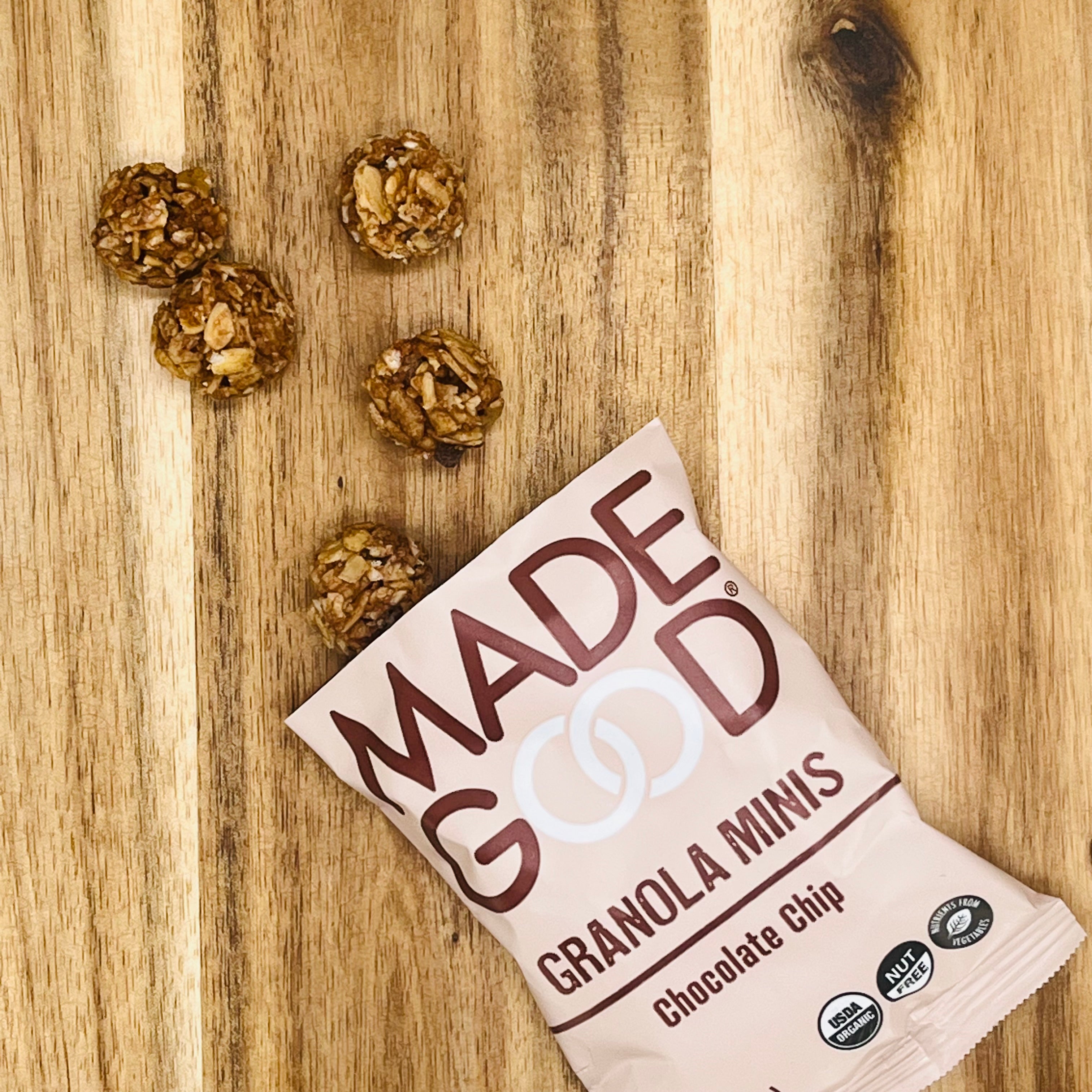 A packet of MadeGood Chocolate Chip Granola Minis (1-0.85 oz Bag) lies on a wooden surface with five granola balls scattered next to it, featuring organic ingredients and gluten-free oats.