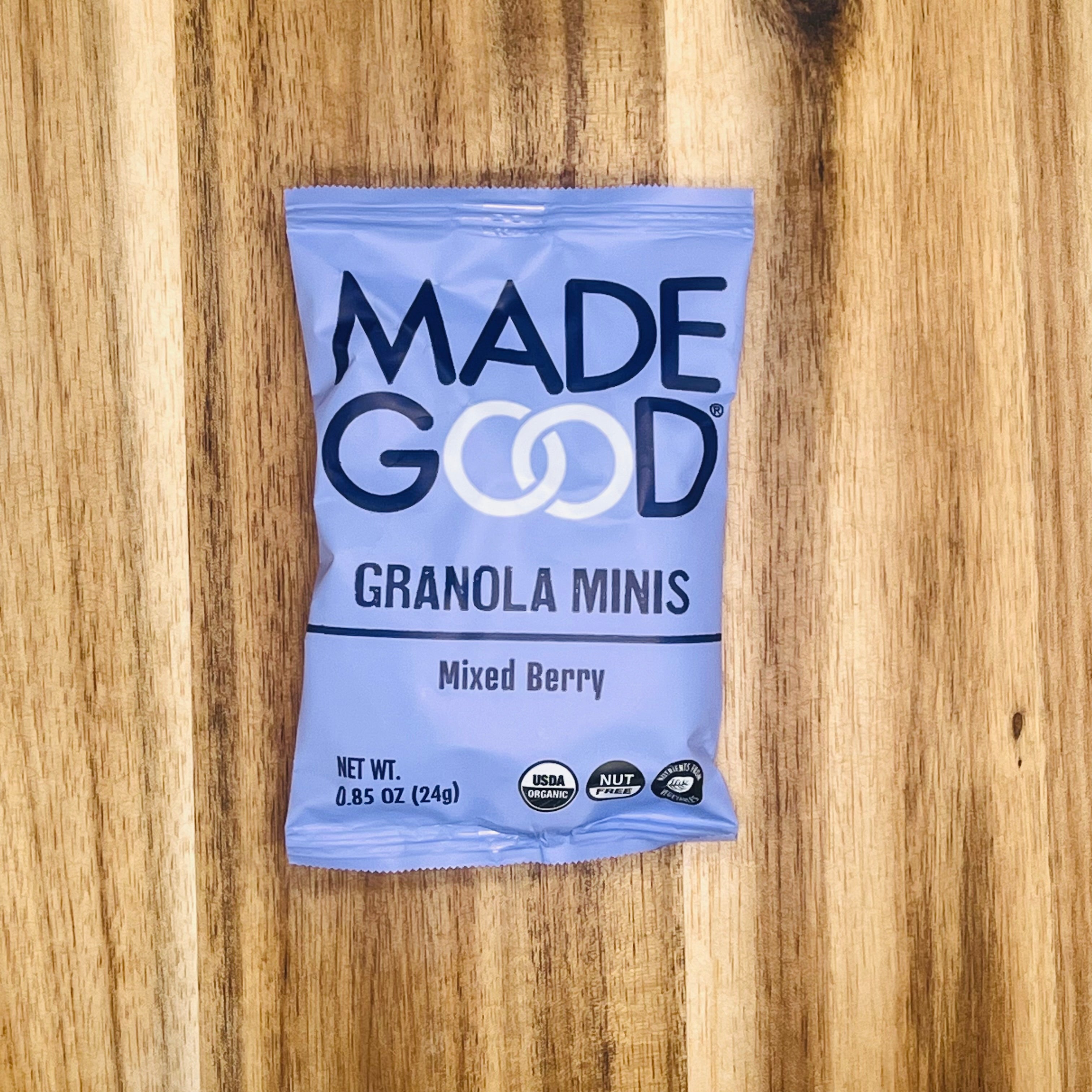 A 0.85 oz (24g) packet of MadeGood Organic Gluten Free Granola Minis in Mixed Berry flavor, displayed on a wooden surface. The purple packaging prominently features the USDA Organic and Nut-Free symbols, emphasizing that it is a nutrient-rich snack suitable for those who require a gluten-free diet.