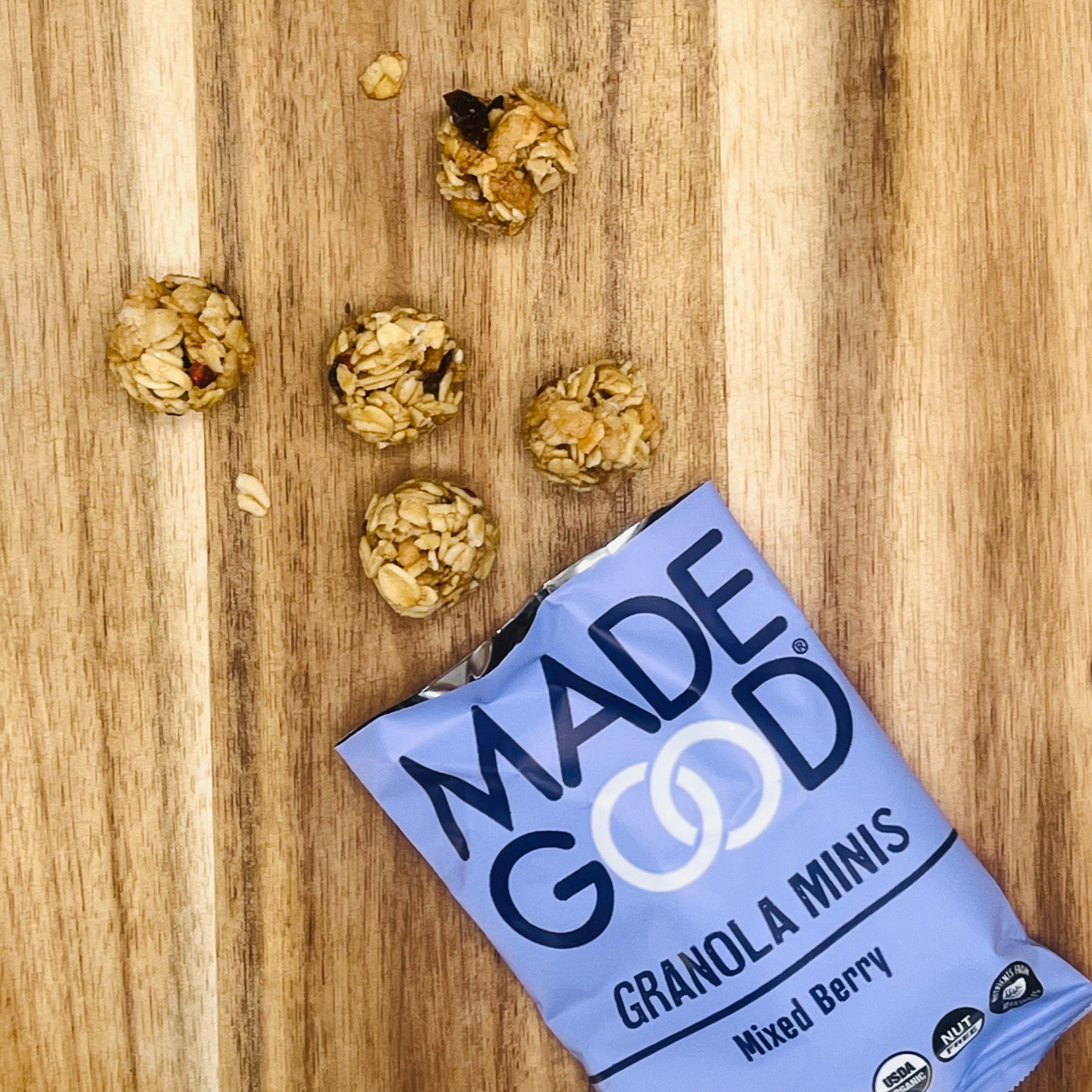 A partially opened MadeGood Organic Gluten Free Granola Minis packet, labeled "Mixed Berry," sits on a wooden surface with some granola balls scattered around. This nutrient-rich snack proudly displays various certifications at the bottom, highlighting its allergy-friendly and gluten-free attributes.