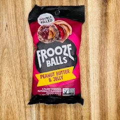 A 2.5 oz package of Frooze Balls Peanut Butter & Jelly from the brand Frooze, double filled with peanut butter and jelly, is placed on a wooden surface. The packaging emphasizes that the product is vegan, plant-based, naturally flavored, gluten-free, and contains no preservatives.