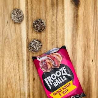 A packet of Frooze Frooze Balls Peanut Butter & Jelly - 2.5 oz is lying open on a wooden surface with three vegan, gluten-free snack balls placed nearby.