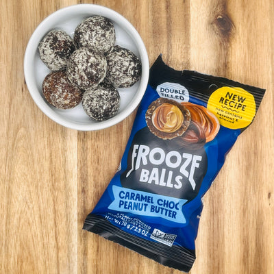 A white bowl with six chocolate-coated balls sits beside a package of Frooze Balls Chocolate Caramel Peanut Butter - Double-Filled Energy Balls 2.5 oz - 1 Count, all arranged on a wooden surface.