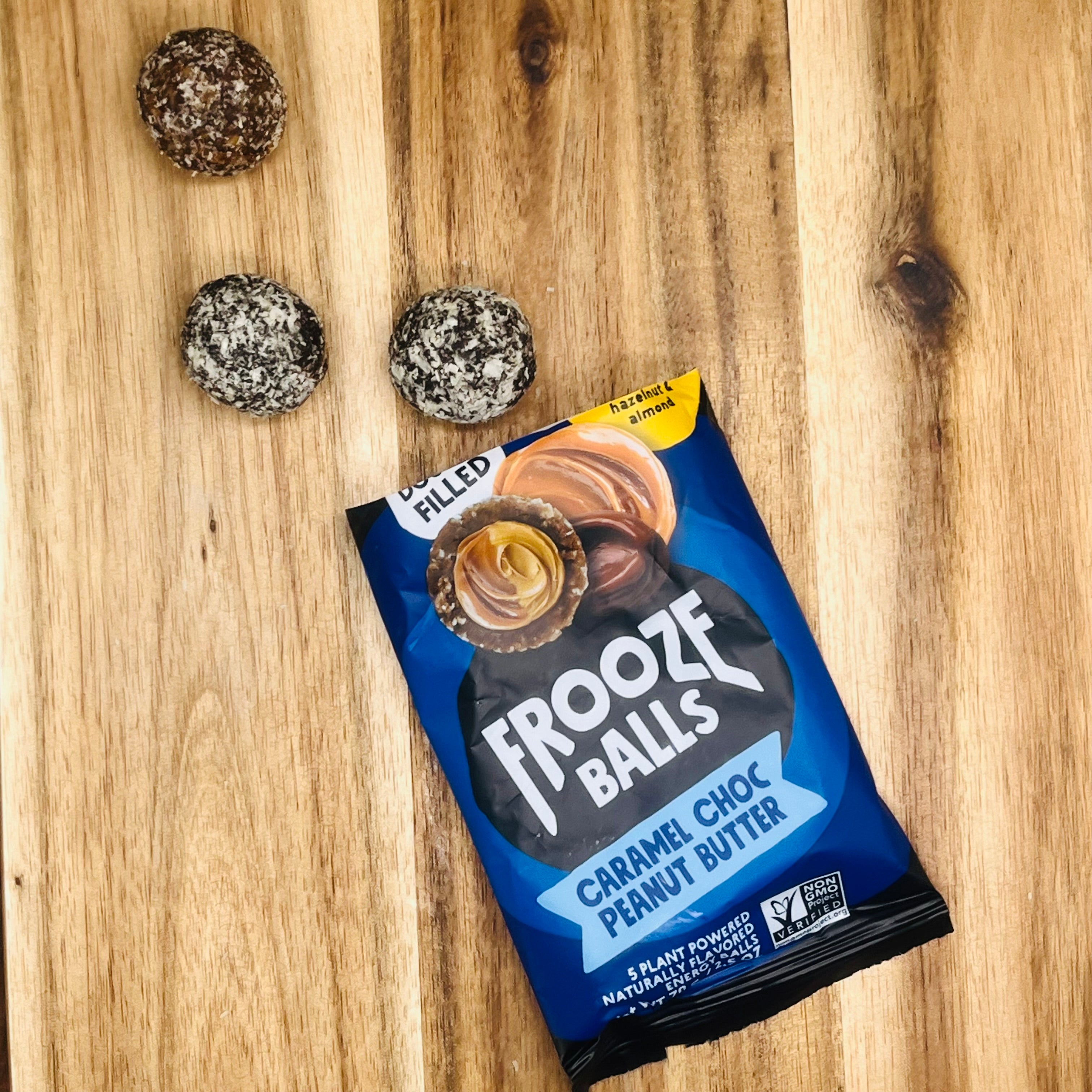 A packet labeled "Frooze Balls Chocolate Caramel Peanut Butter - Double-Filled Energy Balls 2.5 oz - 1 Count" lies on a wooden surface beside four energy balls with a chocolatey coating, highlighting their plant-based and gluten-free nature.