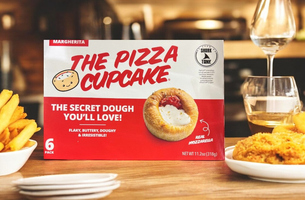 A box of The Pizza Cupcake Margherita-1 Count is on the table with fries and fried chicken. The package displays a cheese and tomato sauce cupcake, highlighting its "Secret Dough You'll Love!" for a classic pizza taste with a unique twist.