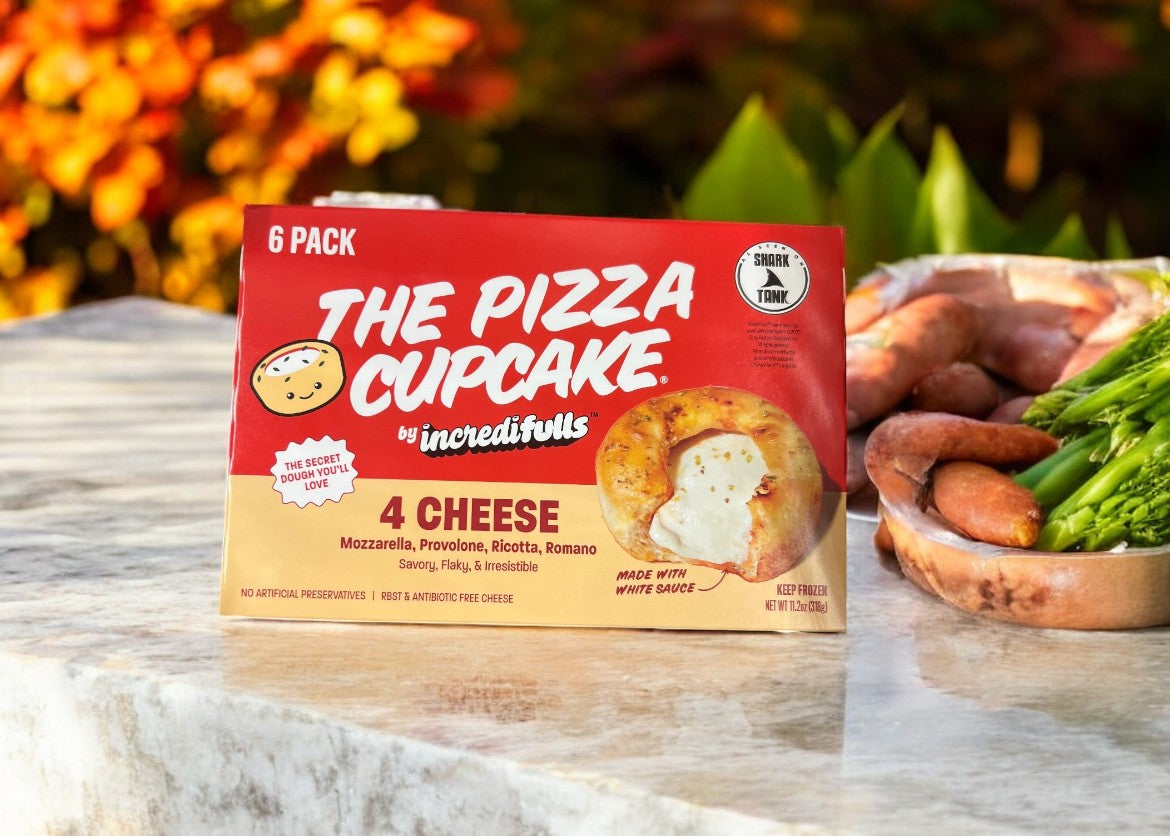 A package of The Pizza Cupcake 4 Cheese - 1 Count, the ultimate cheesy snack from The Pizza Cupcake, is displayed outdoors on a table near greenery and blurred autumn-colored foliage.