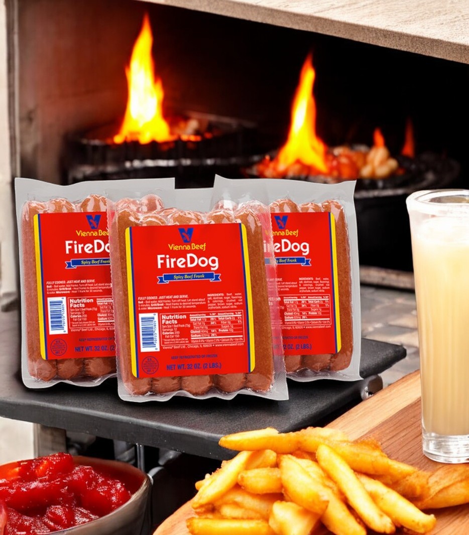 Three packages of Vienna Beef Fire Spicy Jumbo Franks, crafted from premium beef, are displayed by a grill with flames. Fries, a glass of milk, and a small dish of ketchup are in the foreground.